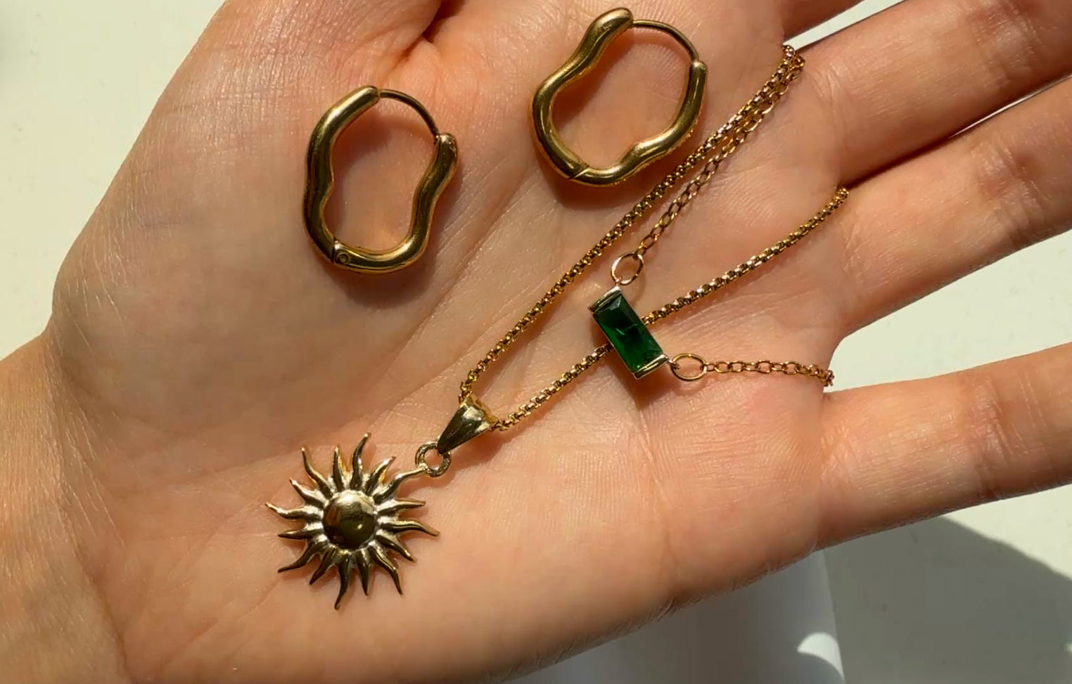 Hand holding gold jewelry with a sun design and green gemstone on a neutral background