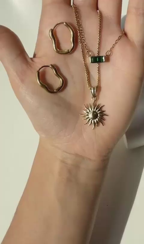 Wavy golden hoop earrings, green rectangle glass necklace, and long sun charm on a golden chain.